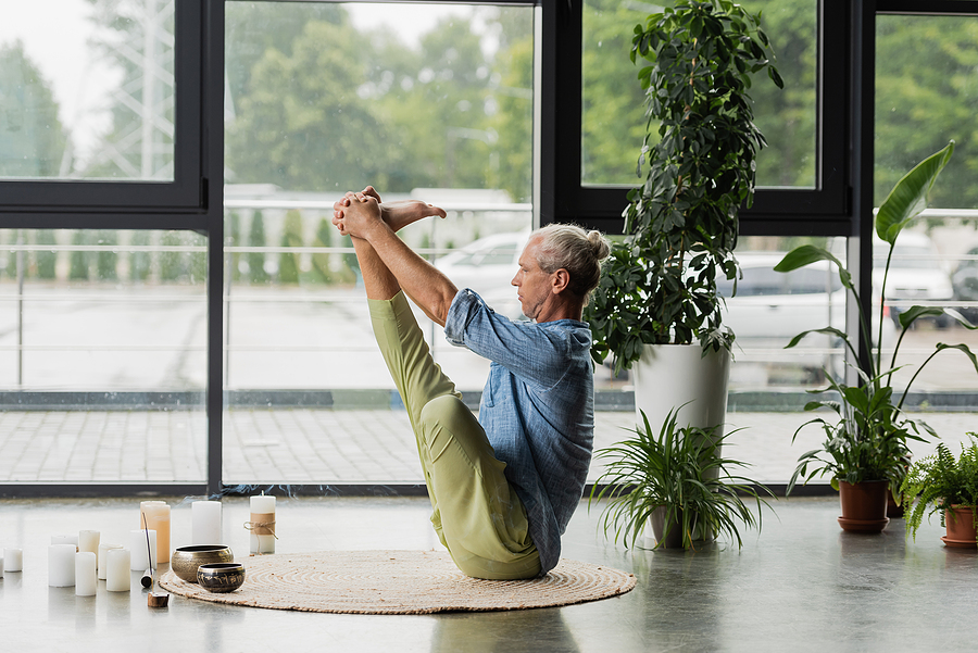 grey haired man doing boat yoga pose near tibetan singing bowls